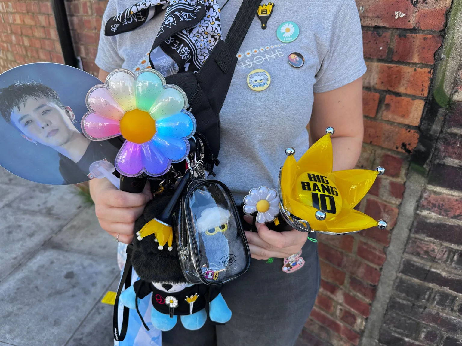 Person holding a collection of concert merchandise and light sticks in the foreground. Items include a flower-shaped light stick with pastel-coloured petals, a yellow crown-shaped light stick marked “BIG BANG 10”, a handheld fan featuring a portrait, and a small flower-shaped accessory. A black cross-body bag is decorated with badges and pins, and holds a clear pouch containing a blue character keyring. The person is standing beside a red brick wall, with a pavement visible to the left.