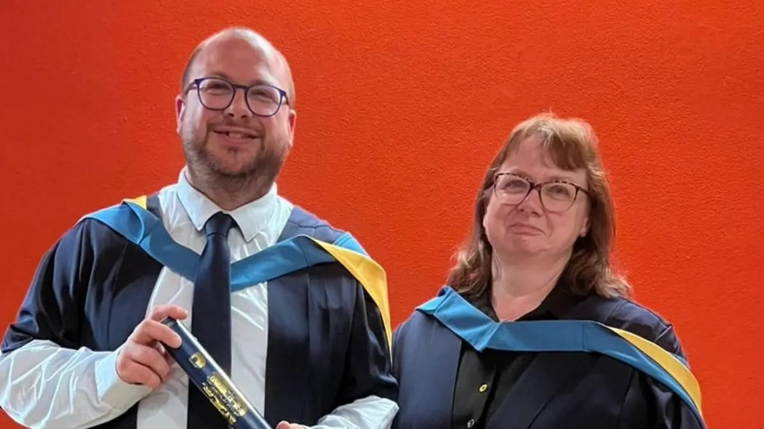 Supplied A man is holding his degree certificate while wearing a blue and yellow graduation robe, blue shirt and blue tie. A woman standing next to him also wears a graduation robe and has a black shirt underneath. They are standing in front of a red background.