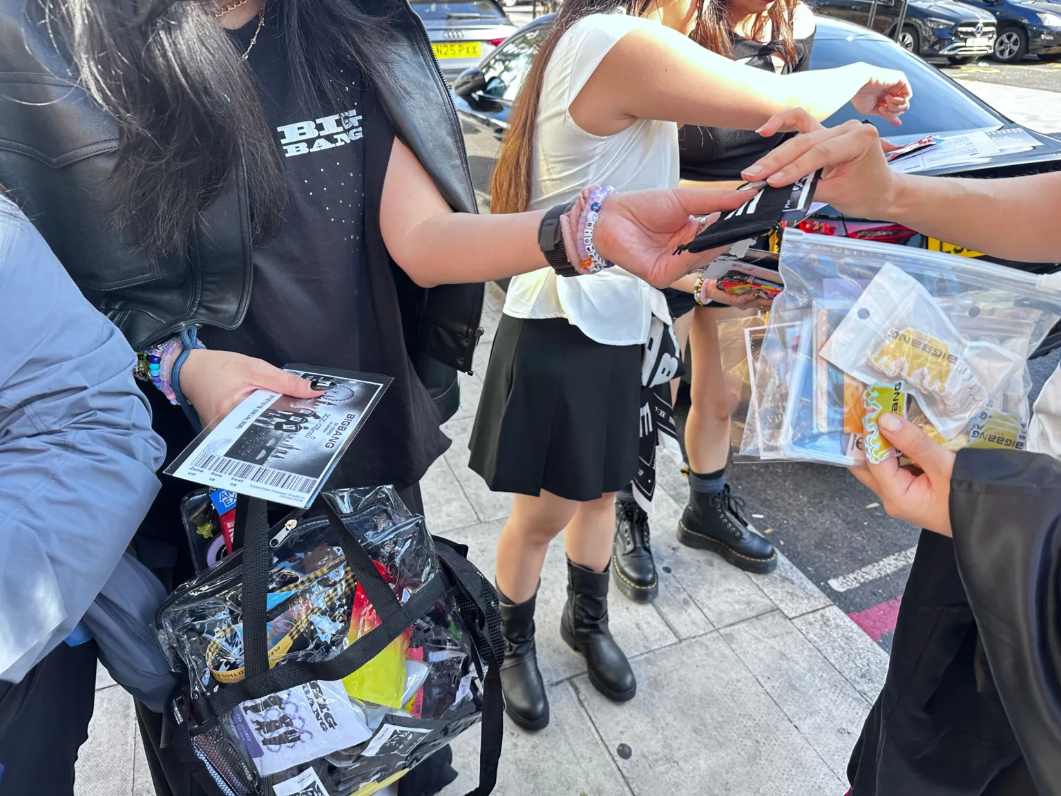 People standing on a pavement exchange small items and wristbands by hand. In the foreground, a clear plastic zip bag contains concert-themed freebies, including stickers, cards and keyrings. Another person holds a large transparent tote bag filled with merchandise and printed cards. Visible clothing includes a black “BIGBANG” T-shirt and a black sleeveless jacket. Parked cars and additional people are in the background beside the kerb.