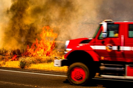 An emergency vehicle driving along a road as flames and smoke rage in the background.