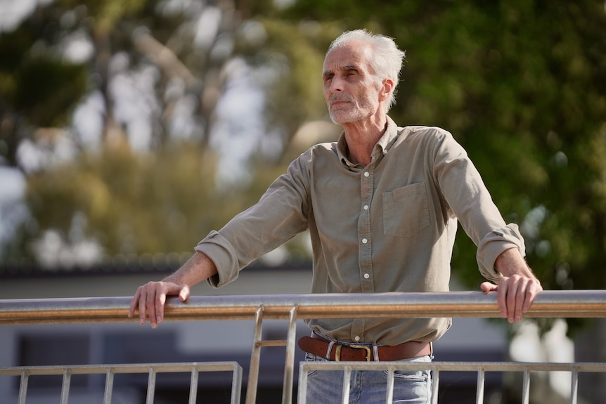 A man with white hair looks over a balcony with trees in the background.