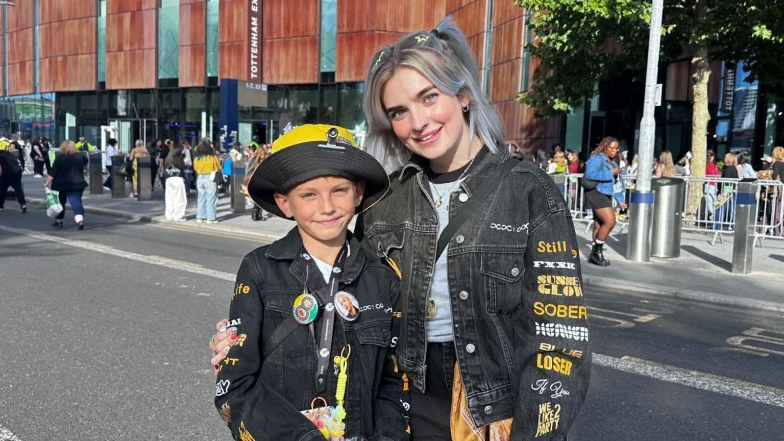 Mum and son standing in the foreground on a road outside a stadium. Both are wearing black denim jackets decorated with printed text, badges and pins. In the background, queues of people stand behind metal barriers outside the stadium entrance, with trees, bollards and signage visible along the pavement.