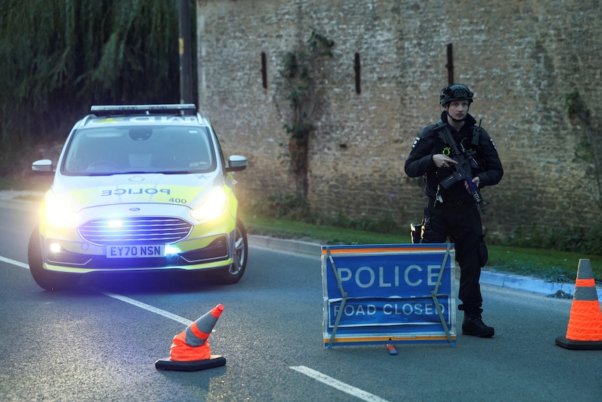 An armed police officer stands on the road in front of a police car at dawn on rural road