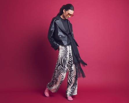 A model wears zebra-print trousers with black leather jacket, scarf, sunglasses and pink mules, all against a maroon studio backdrop