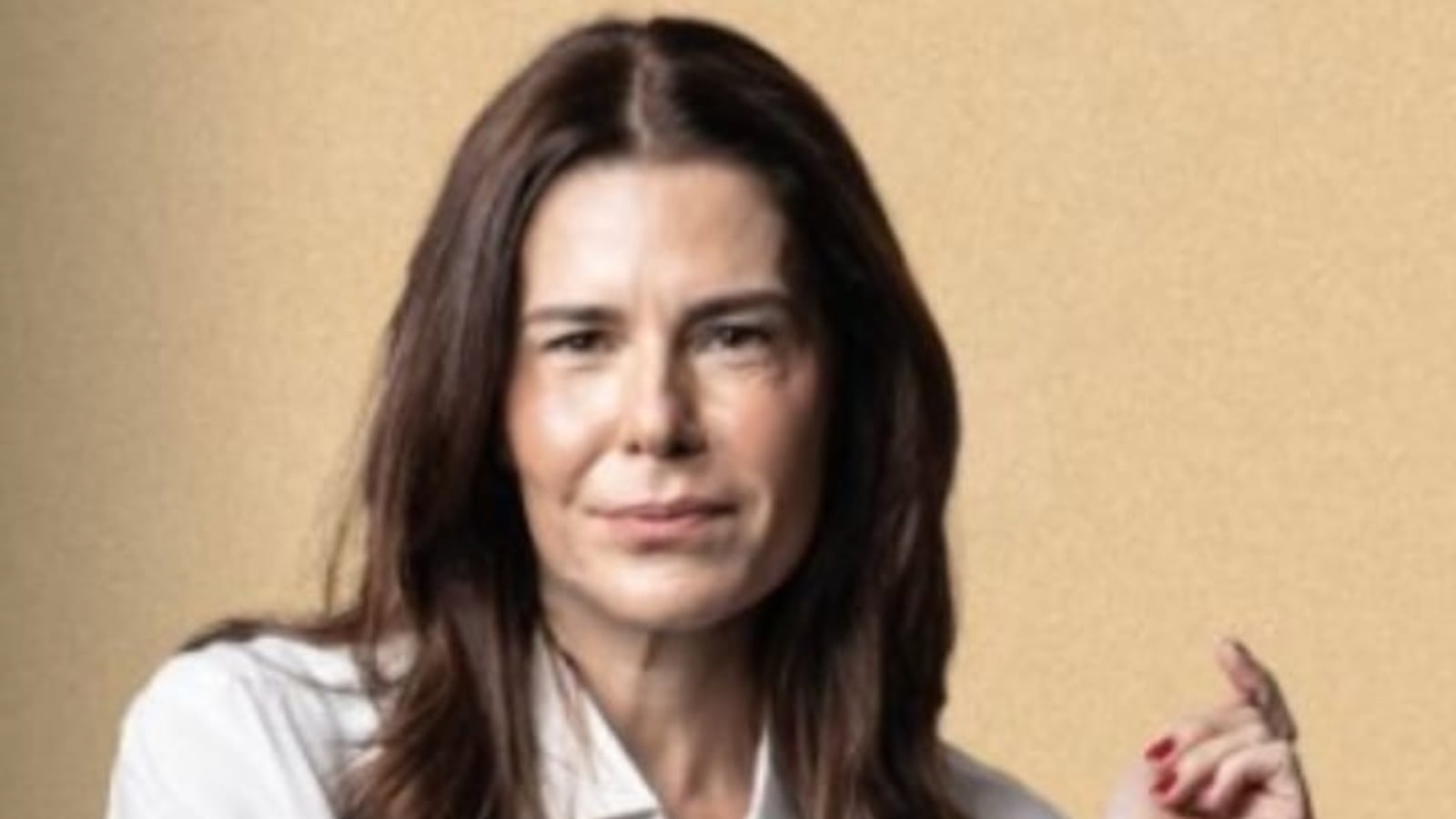 Tati Oliva, uma mulher com cabelos longos e castanhos, usando uma camisa branca, está em pé com os braços cruzados. Ela tem uma expressão séria e segura um cigarro entre os dedos da mão direita. O fundo da imagem é de uma cor bege clara, criando um contraste suave com a roupa dela.